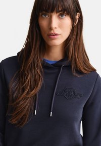 Mladi žena s dugom smeđom kosom u tamnoplavom duksu s kapuljačom i logom GANT te vezicama, gleda prema naprijed na jednobojnoj pozadini.