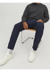 Námořnické chinos kalhoty s volnějším střihem, doplněné šedým tričkem s dlouhým rukávem. Bílé tenisky, viditelné ponožky s logem, sedící na dřevěné židli.