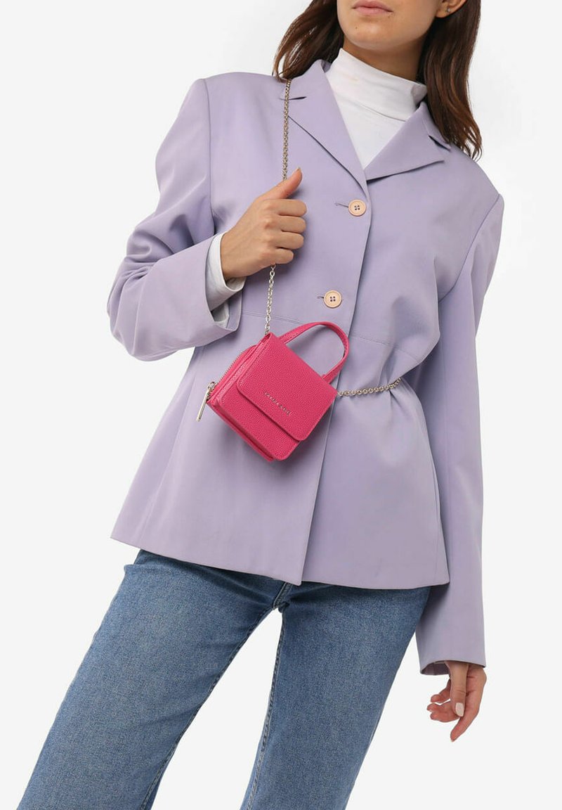 Kleine roze leren handtas met een gestructureerde afwerking, rechthoekige vorm, bovenhandvat en kettingriem, gedragen bij een lila blazer.