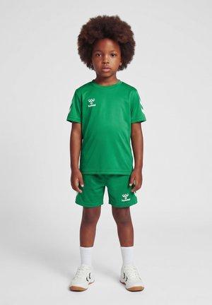 Barn med afro-hår iført en grøn sportstøj, hvide sokker og hvide sneakers, stående mod en ensfarvet lys baggrund.