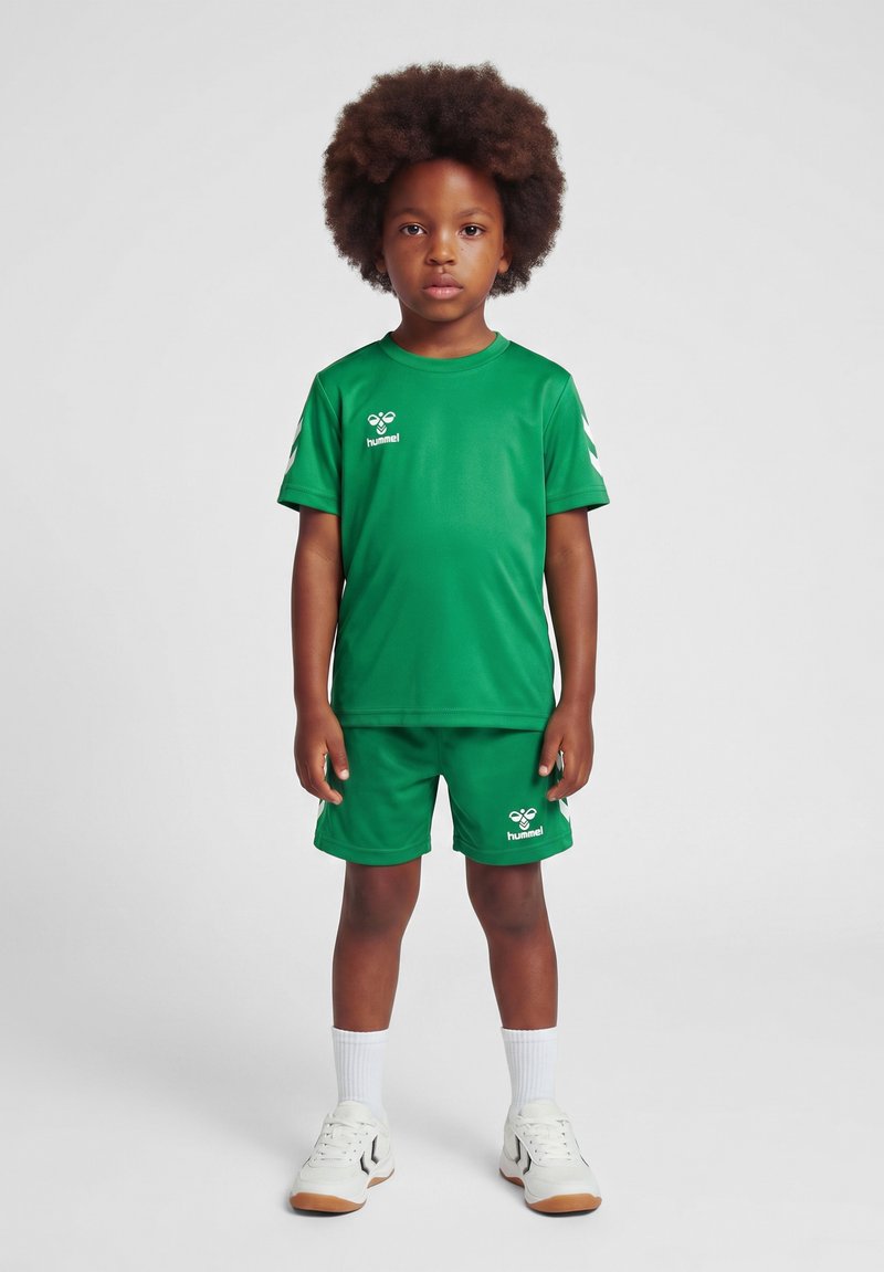 Kind met afro haar, gekleed in een groen sportoutfit, witte sokken en witte sneakers, staand tegen een effen lichte achtergrond.