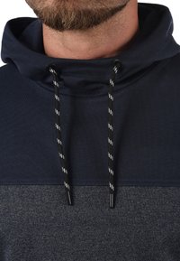 Felpa blu navy con una sezione inferiore grigia tessurizzata, caratterizzata da un colletto alto, lacci neri e bianchi e una finitura in tessuto liscio.