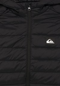 Black quilted jacket with central zipper and small rectangular patch showing a white mountain wave logo on the chest.