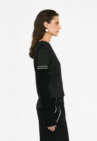 Maglione nero con dettagli in filo metallico, caratterizzato da una finitura testurizzata, maniche corte e maniche lunghe a contrasto. Orecchini a forma di cuore di rilievo.