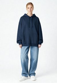 Tengerészkék kapucnis pulóver kenguruzsebbel, világoskék bő szárú farmerral és fehér tornacipővel párosítva. Simaságot és puhaságot sugárzó anyag.