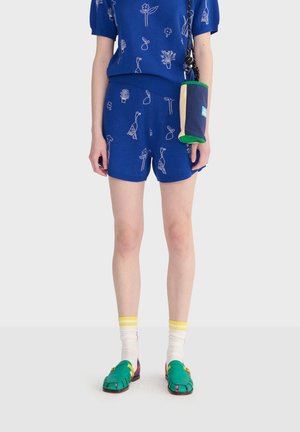 Personne portant un short et un haut bleus avec des motifs blancs de plantes et d'oiseaux, des chaussettes blanches à rayures jaunes, des chaussures vertes et un petit sac bandoulière bleu.