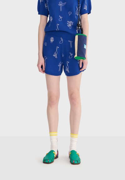 Persona vistiendo pantalones cortos y camiseta azul con patrones blancos de plantas y aves, calcetines blancos con rayas amarillas, zapatos verdes y una pequeña bolsa cruzada azul.