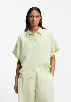 Femeie cu păr lung și închis la culoare, purtând o cămașă verde deschis cu mâneci scurte și nasturi, și pantaloni largi asortați, stând cu o mână în buzunar.