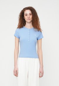 Γαλάζιο ριμπ κοντομάνικο τοπ με κούμπωμα στο ντεκολτέ και κεντημένο λογότυπο στο στήθος, συμπληρωμένο από εφαρμοστή σχεδίαση.