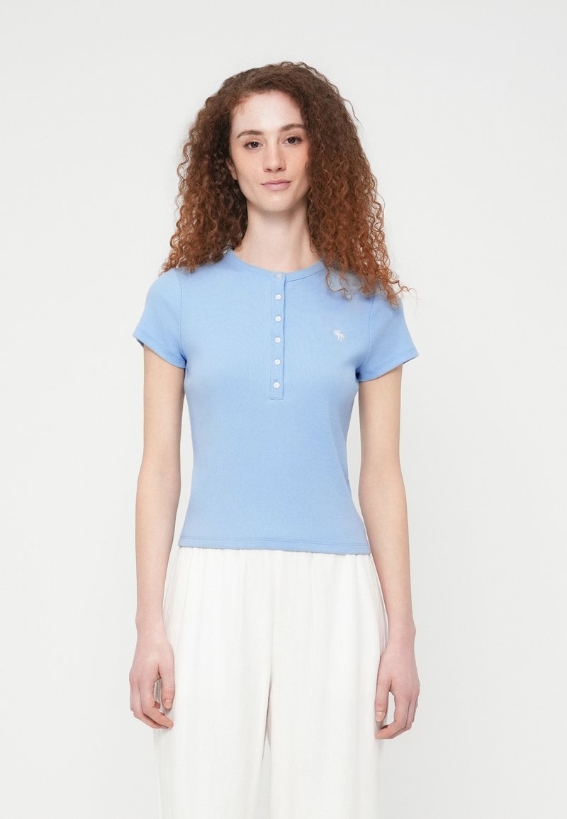 Γαλάζιο ριμπ κοντομάνικο τοπ με κούμπωμα στο ντεκολτέ και κεντημένο λογότυπο στο στήθος, συμπληρωμένο από εφαρμοστή σχεδίαση.