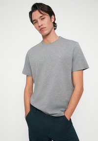 Jaunas vyras su tamsiais plaukais, vilkintis pilką trumpą rankovę marškinėlius ir tamsias kelnes, stovi rankomis kišenėse prieš paprastą foną.