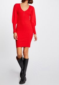 Robe en maille côtelée rouge avec un décolleté en V profond, des manches longues bouffantes et des boutons décoratifs le long du devant. Associée à des bottes hautes noires.