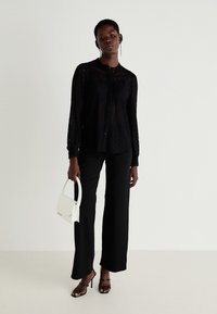 Blouse noire en dentelle avec boutons et manches longues, assortie à un pantalon noir à jambes larges et des talons aiguilles noirs à lanières. Accessoirisée avec un sac à main blanc.