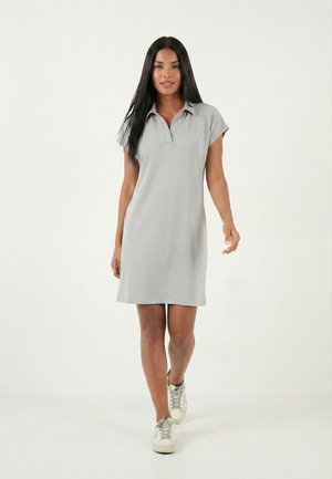 Mujer caminando hacia adelante, vestida con un vestido tipo polo de manga corta gris claro y zapatillas blancas, sobre un fondo blanco liso.