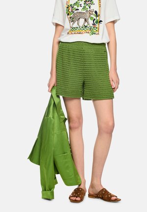 Frau trägt grüne Häkel-Shorts, weißes T-Shirt mit Leopardenmuster, hält grüne Jacke und braune Slip-On-Sandalen mit goldenen Ösen.