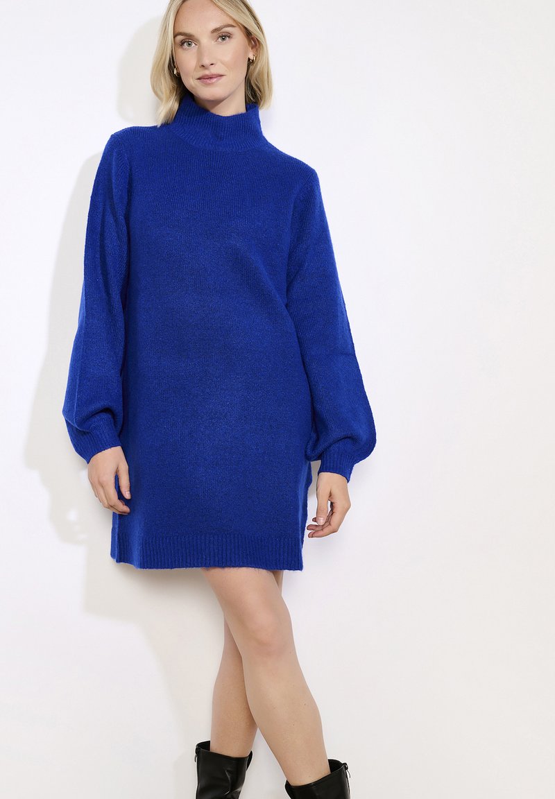 Moteris vilkinti kobalto mėlynumo megztą golfo suknelę ir juodus vidutinio ilgio batus, stovinti prieš paprastą baltą foną.