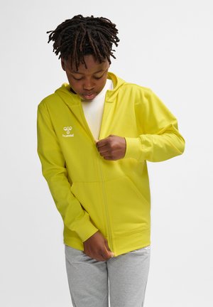 Gul zip-up hættejakke, blødt stof, kængurulommer, hvidt logo på brystet, parret med grå sweatpants. Teksturen fremstår glat.