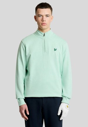Ung mand iført en lysegrøn quarter-zip trøje med et lille fuglelogo og en hvid golfhandske på højre hånd.
