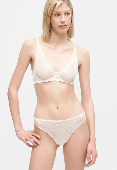 Γυναίκα που φορά διαφανές λευκό σουτιέν και αντίστοιχο εσώρουχο με διακριτικά γεωμετρικά λογότυπα, στέκεται με το ένα χέρι στη μέση μπροστά σε απλό φόντο.