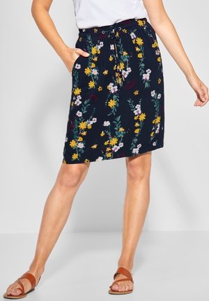 Frau trägt einen knieumspielenden marineblauen Rock mit gelben und weißen Blumenmustern, ein weißes Oberteil und braune flache Sandalen.