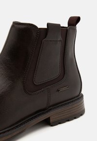 Hnědé kožené kotníkové boty s elastickými bočními panely, robustní podešví a texturovaným povrchem. Obsahují dekorativní prošívání a detaily s logem.