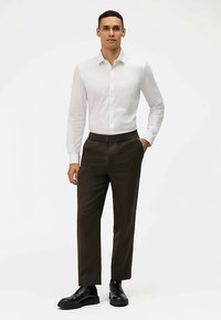 Weißes Langarmhemd mit Knopfleiste, kombiniert mit dunkelbraunen Hosen mit elastischem Bund und schwarzen Schnürschuhen. Glatte Stoffstruktur.