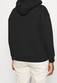 Sudadera negra con capucha ajustable, puños y dobladillo de canalé, textura suave. Lleva puesta sobre pantalones de color claro, vista desde la parte de atrás.