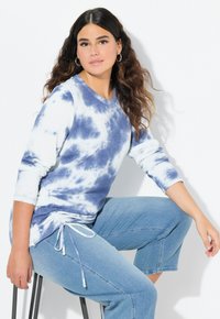 Sudadera de manga larga teñida en azul y blanco, con un corte holgado y detalle de cordón en el dobladillo, combinada con jeans de pierna ancha en azul claro.