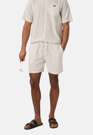 Mężczyzna noszący beżową koszulę w paski z krótkim rękawem i dopasowane szorty, trzymający okulary przeciwsłoneczne w jednej ręce, noszący czarne sandały.