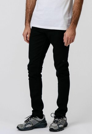 Muž oblečený v čiernych slim fit nohaviciach, bielom tričku a sivých bežeckých topánkach, stojaci pred jednofarebným svetlým pozadím.