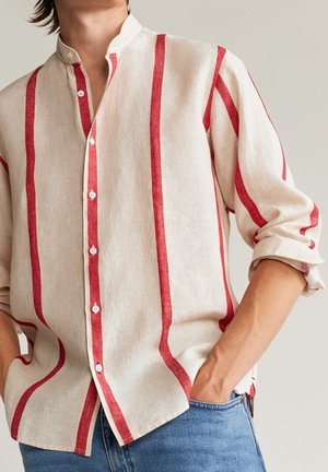 Personne portant une chemise beige à boutons avec des rayures rouges verticales et des manches retroussées, avec une main dans la poche d'un jean bleu.