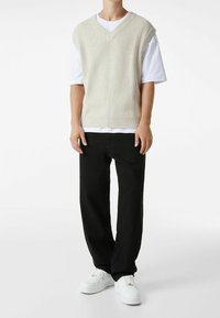 Suéter sin mangas de punto color beige claro con cuello en V y textura acanalada, llevado sobre una camiseta blanca, combinado con pantalones negros y zapatillas blancas.