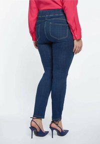 Kapeamattomasti siniset farkut venyvällä vyötäröosalla, joissa on kaksi takataskua ja keltainen tikkaus, yhdistettynä sinisiin korkokenkiin.