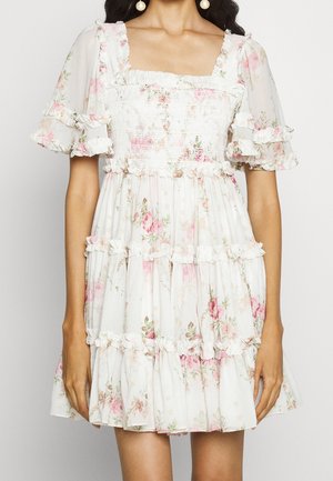 Vrouw die een korte witte jurk draagt met roze bloemenprint, pofmouwen, een geplooide taille en gelaagde ruches.