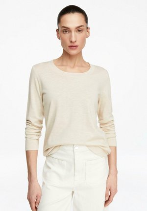 Kobieta z upiętymi włosami, ubrana w beżową bluzkę z długim rękawem i wysokim stanem ecru spodnie na jednolitym białym tle.