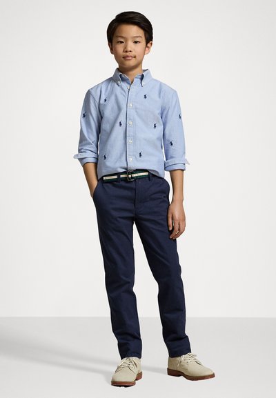 Camicia azzurro chiaro con bottoni e loghi ricamati, chinos blu navy e scarpe beige. Presenta una cintura multi-strisce e maniche arrotolate.