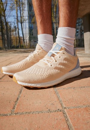 Person trägt beige und hellblaue Adidas Laufschuhe mit weißen Socken und steht auf einem gepflasterten Gehweg im Freien.