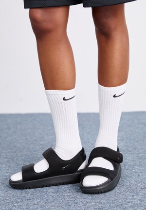 Benens met witte Nike sportsokken en zwarte Nike sandalen met verstelbare bandjes op een blauw tapijt.