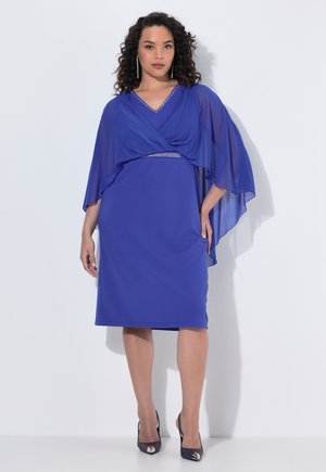 Frau mit lockigem Haar, die ein knielanges blaues Kleid mit transparenten Umhangärmeln und dunklen High Heels trägt, steht vor einem weißen Hintergrund.