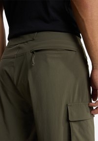 Un pantalon cargo vert olive doté d'une poche arrière zippée et d'une poche latérale, fabriqué en tissu léger avec une texture lisse.