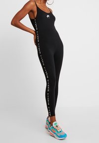 Svart ärmlös jumpsuit med en åtsittande design, med vit "Nike Air" branding längs sidorna. Matchas med färgglada sneakers.