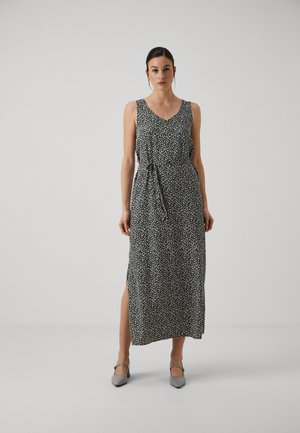 Robe maxi noire avec un motif floral blanc, décolleté en V, sans manches, taille nouée, fente latérale. Associée à des chaussures gris clair. Texture de tissu lisse.