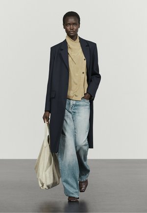 Femme portant un long manteau foncé, une blouse beige, un jean large bleu clair, des chaussures marron et portant un grand sac beige, marchant sur un sol gris.