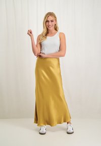 Gele satijnen maxi-jurk met een gladde textuur, gecombineerd met een witte geribbelde tanktop en witte sneakers. Model poseert met één hand omhoog.