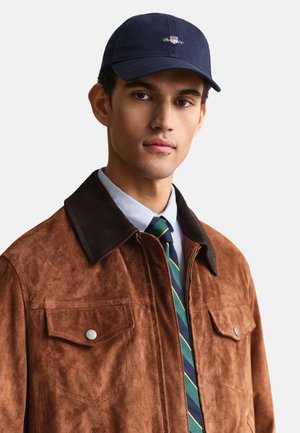 Homme portant une veste en daim marron avec un col foncé, une chemise bleue, une cravate rayée verte et bleue, et une casquette bleu marine avec un petit logo.