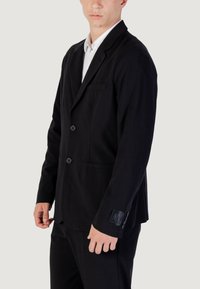 Uomo che indossa un blazer nero a due bottoni sopra una camicia bianca, in piedi contro uno sfondo chiaro uniforme, con il logo AX visibile sul polsino.