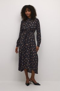 Navyblaues Hemdkleid mit beigen Punkten, an der Taille gebunden. Lange Ärmel, fließender Stoff und ein leicht verlängertes Rückenteil. Schwarze High Heels.