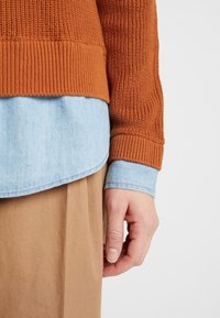 Ruosteenvärinen neulottu villapaita, jossa on resorireuna, kerrospukeutuneena vaaleansinisen farkkupaidan päälle, kädellä pidettynä vaaleanruskeaa kangasta vasten.