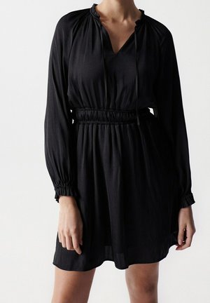 Femme portant une robe noire à manches longues avec taille et poignets élastiqués froncés, debout devant un fond blanc uni.