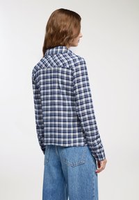 Camicia a quadri nei colori blu navy, bianco e azzurro chiaro; presenta maniche lunghe, colletto e un orlo posteriore dritto. Indossata con jeans azzurri chiari.